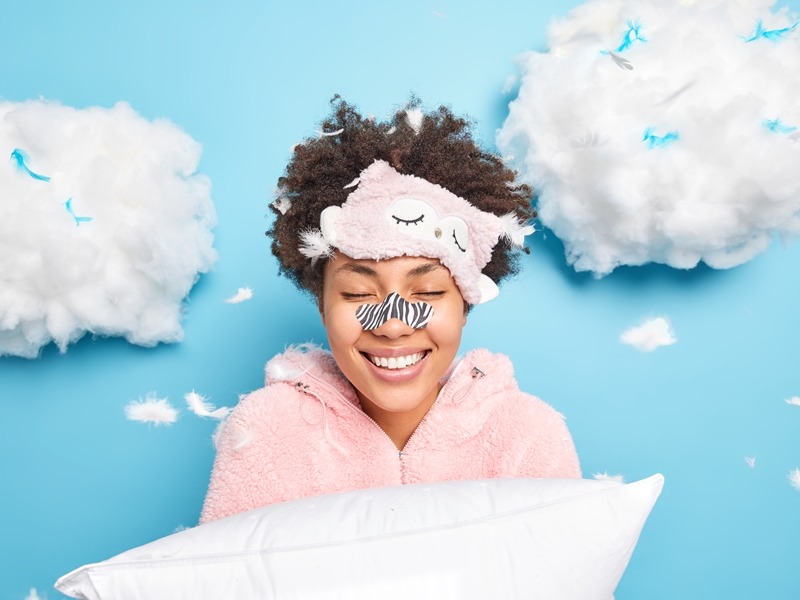 Happy curly Afro American woman gets rid of black dotes on nose using special patch wants to have flawless clean skin smiles gently dressed in pajama holds pillow poses around flying feathers